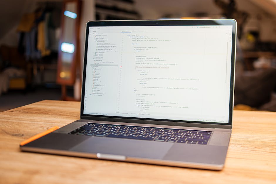 A close-up of a laptop open on a wooden desk displaying a coding interface with lines of colorful code, accompanied by a white smartphone placed beside it. The scene suggests an indoor workspace envir